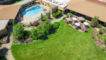 An aerial view shows a pool area with lounge chairs and umbrellas, adjacent to a manicured lawn and garden with patio tables and more umbrellas.