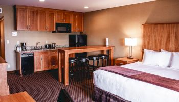 The image shows a hotel room with a bed, a kitchenette, a small dining area, and a desk with a chair.