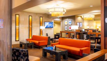 A modern lounge area with orange sofas, black tables, and contemporary decor under warm lighting, featuring a wall-mounted TV and framed art.