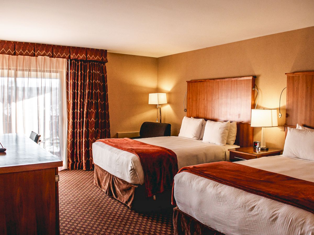 The image shows a hotel room with two double beds, a TV, a desk, and a lamp by the window. The room has warm lighting and earth-toned decor.