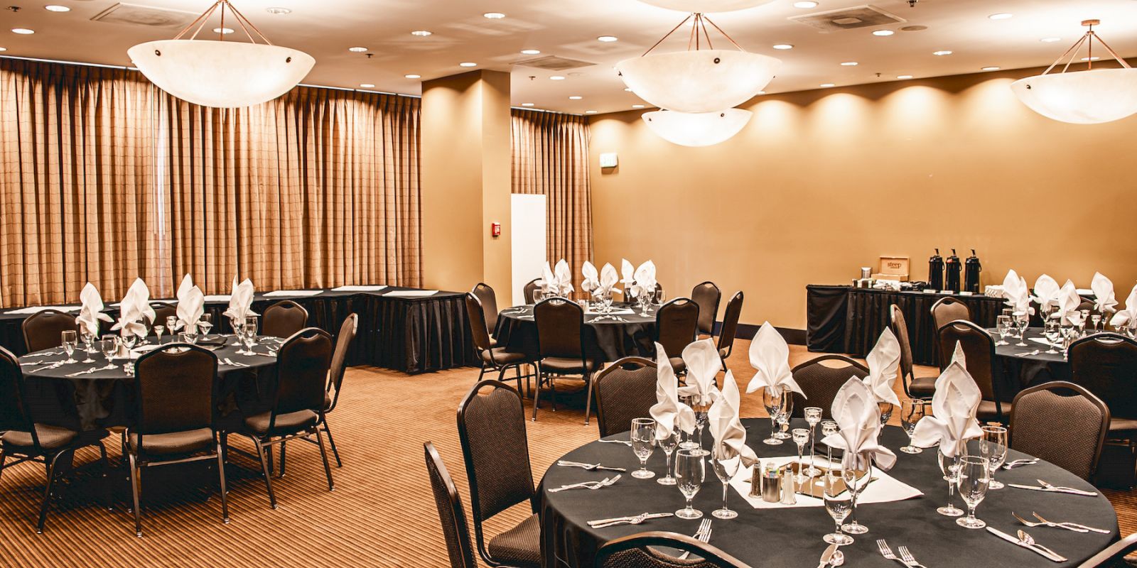 A banquet hall is set with round tables, each adorned with folded napkins, wine glasses, and silverware, lit by ceiling lights.