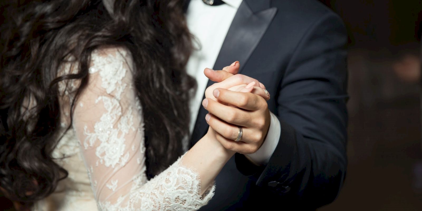 A couple is dressed formally, dancing together. The woman is wearing a lace wedding dress, and the man is in a suit and tie, holding hands.