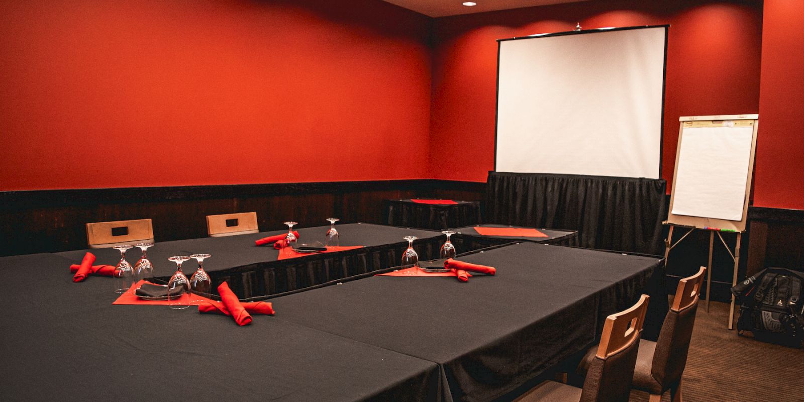 A conference room with a U-shaped table arrangement, black tablecloths, red napkins, wine glasses, a screen, and a flipchart.