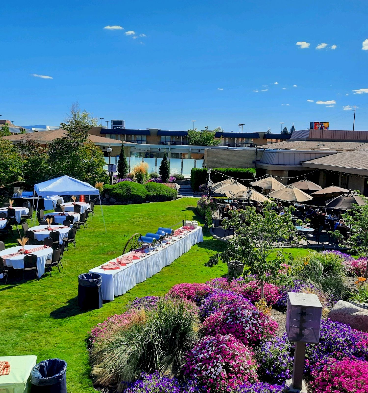 The image shows an outdoor event setup with tables, chairs, and tents on a lawn surrounded by colorful flowers, under a clear blue sky.