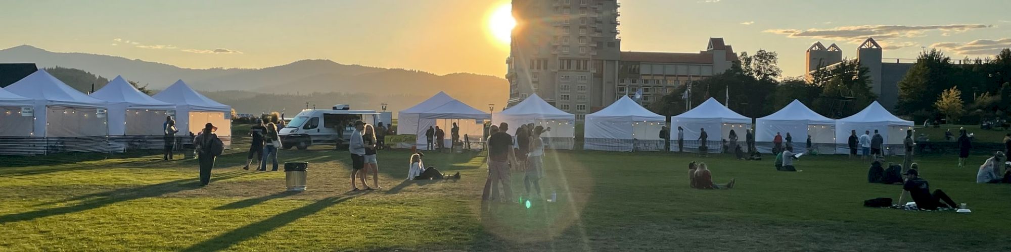 This image shows a grassy field with people, white tents, and a setting sun behind a tall building in the distance.