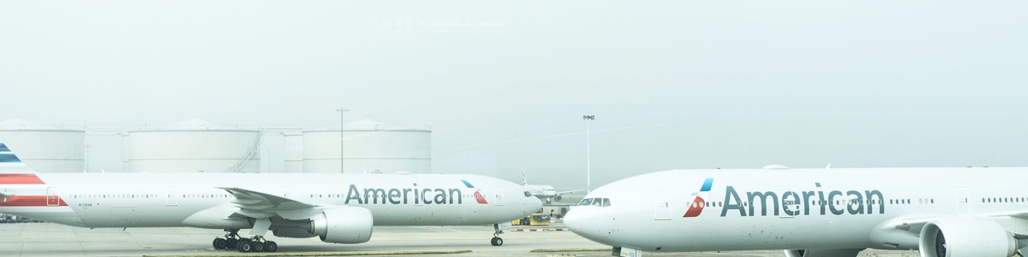 Two American Airlines planes are parked at an airport with fuel storage tanks in the background and ground vehicles in the foreground.