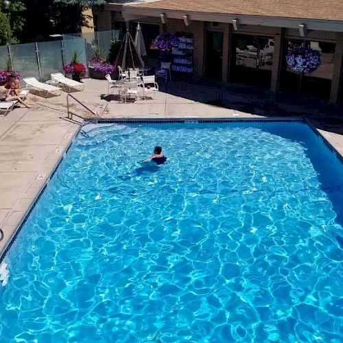 A large outdoor swimming pool with clear blue water, surrounding lounge chairs, and umbrellas. One person is swimming in the pool at the center.