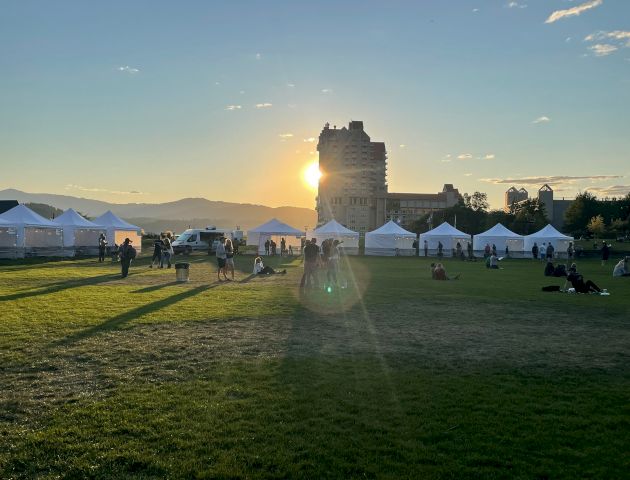 A park scene at sunset with white tents set up, people walking and sitting, and a building in the background, mountain landscape visible.