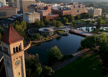 The image shows a cityscape with a prominent clock tower, surrounded by buildings, trees, a body of water, and a park area with pathways.