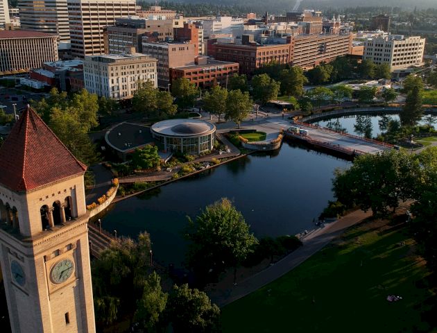 The image shows a cityscape with a prominent clock tower, surrounded by buildings, trees, a body of water, and a park area with pathways.