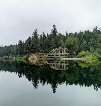 A serene scene of a forested area with a bridge reflected on the calm water of a lake or river.