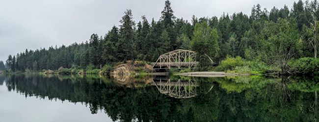A serene scene of a forested area with a bridge reflected on the calm water of a lake or river.