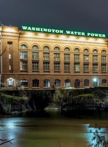A night view of a brick building with "WASHINGTON WATER POWER" in green lights, situated by a river with trees and other buildings surrounding it.