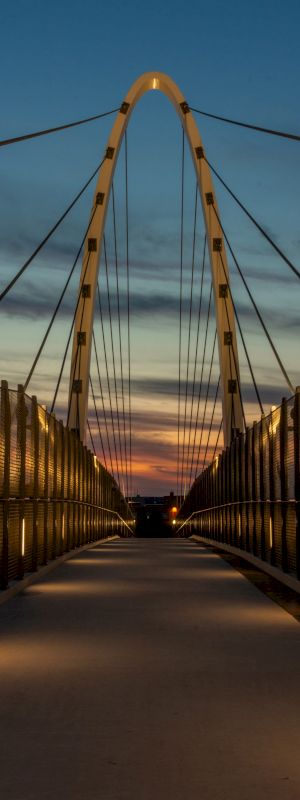 A beautifully lit pedestrian bridge at dusk, with a dramatic sky in the background and modern architectural lines.