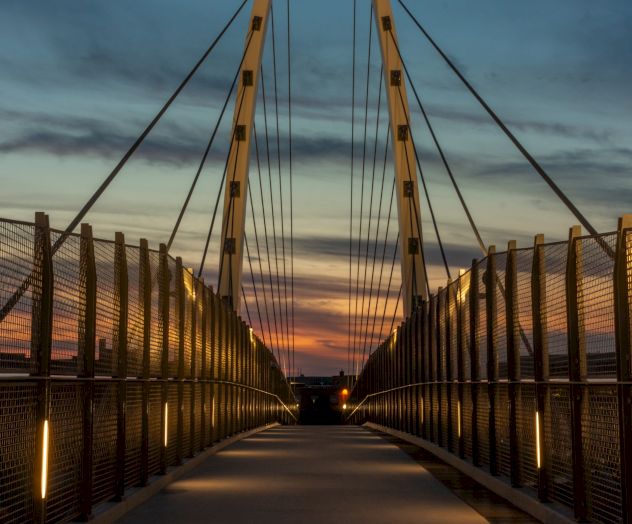 A beautifully lit pedestrian bridge at dusk, with a dramatic sky in the background and modern architectural lines.