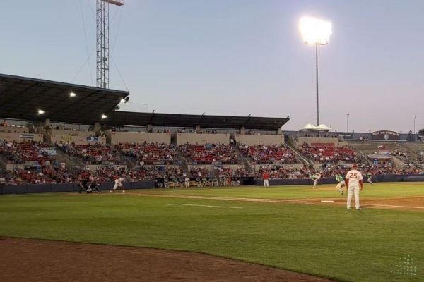 A baseball game is taking place at a well-lit stadium in the evening, with players on the field and a crowd watching from the stands.