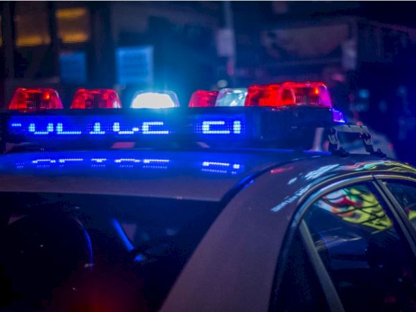 The image shows a police car with its red and blue lights flashing in the night, reflecting the word "POLICE" in blue on the side, indicating active duty.