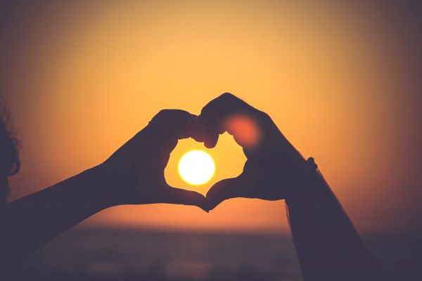 The image shows hands forming a heart shape around a setting sun, creating a romantic or loving mood against a sunset background.