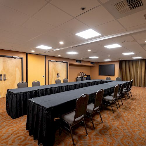 A conference room with a long table, chairs, and a projector screen, featuring carpet flooring and recessed lighting in a neutral decor.