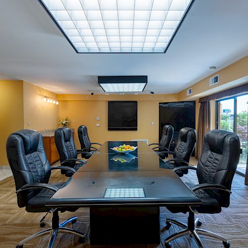 A conference room with a long table, black chairs, a large ceiling light, and a bowl of fruit in the center; windows are on one side.