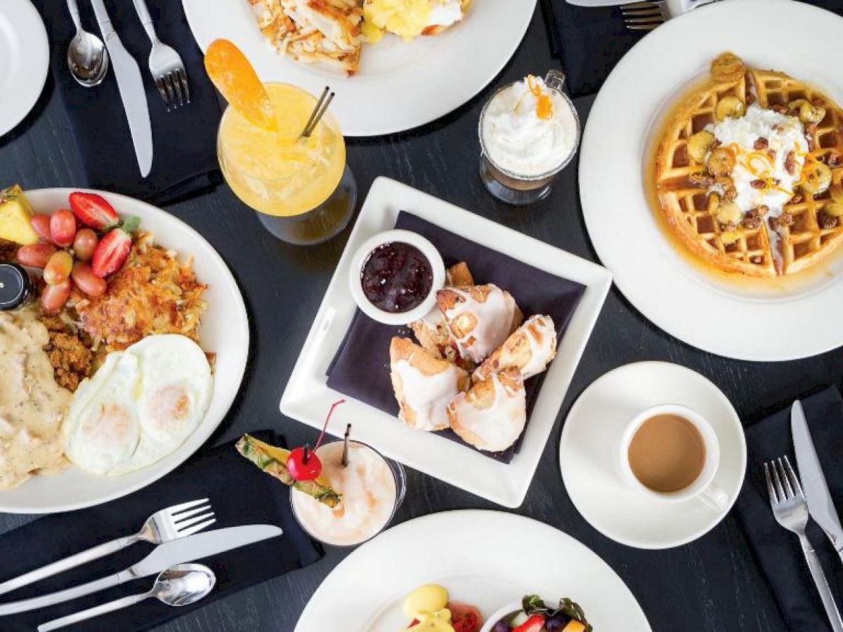 A table set with various breakfast dishes, including eggs, waffles, fruit, drinks, and pastries. Cutlery and napkins are also present.