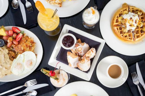 An overhead view of a breakfast spread with waffles, eggs, toast, fruit, coffee, and a smoothie on a dark table set with cutlery and napkins.