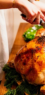 A person is carving a roasted turkey on a wooden table, surrounded by fresh herbs and dishes, suggesting a festive meal preparation.
