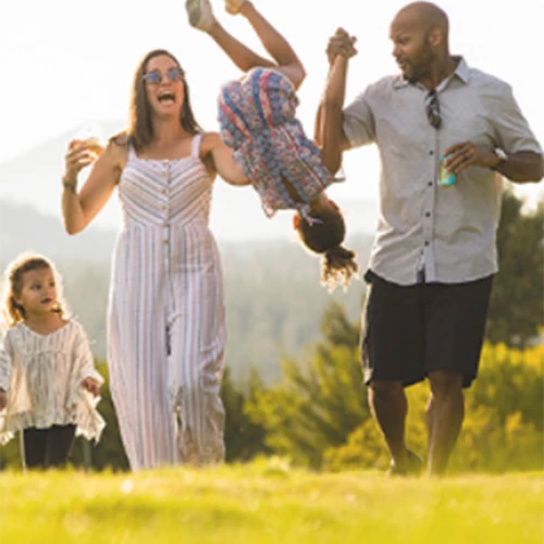 A family is enjoying a sunny day outdoors. An adult man and woman swing a young girl by her hands, while another girl walks beside them.