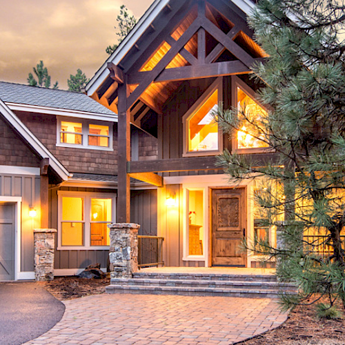 A large, rustic-style house with a stone pathway, three-car garage, and well-lit windows surrounded by tall trees and a dusk sky.