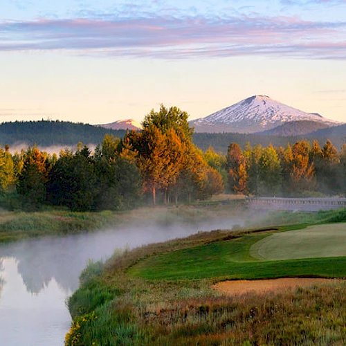 A serene landscape with a river, golf course, lush trees in autumn colors, and a snow-capped mountain in the background during sunrise.