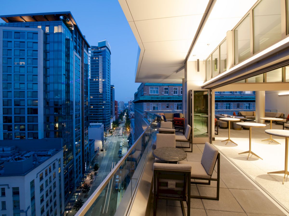 A modern balcony with seating overlooks a city street and skyscrapers at dusk, featuring an indoor-outdoor space with contemporary decor.