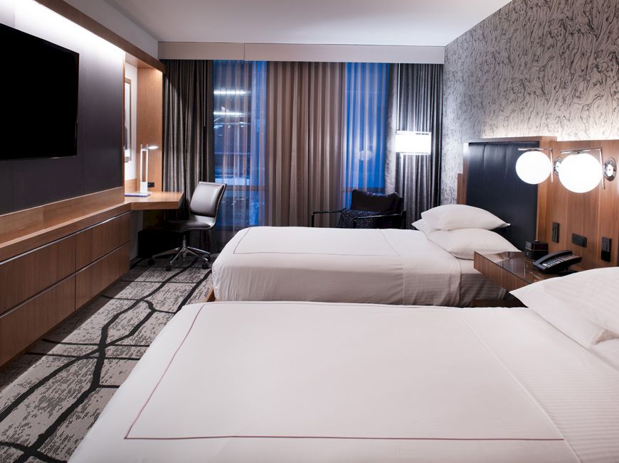 This image shows a hotel room with two neatly made beds, a wall-mounted TV, a work desk, and modern decor.