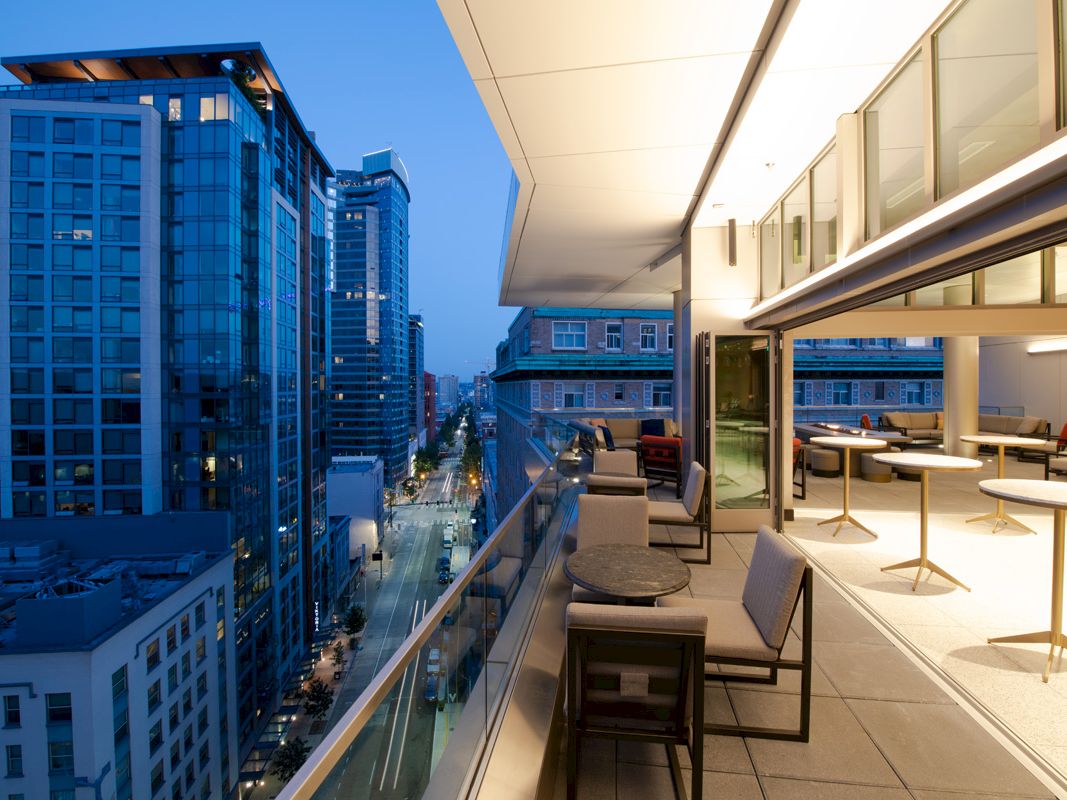 A modern high-rise balcony overlooking a cityscape at dusk, with tables and chairs arranged along the railing.