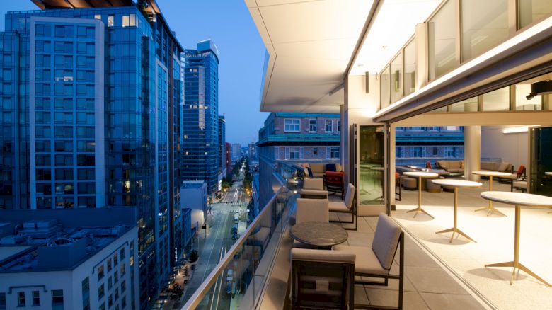The image shows a modern cityscape with tall buildings at dusk, featuring an outdoor balcony with tables, chairs, and a view of the streets below.