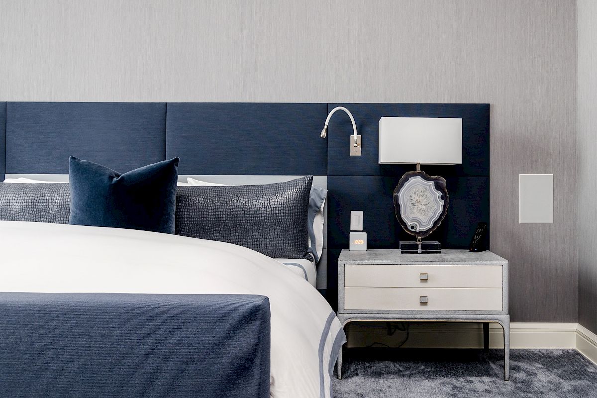 A modern bedroom featuring a blue headboard, white bed linens, a nightstand with a lamp, and decorative pillows in shades of blue and gray.