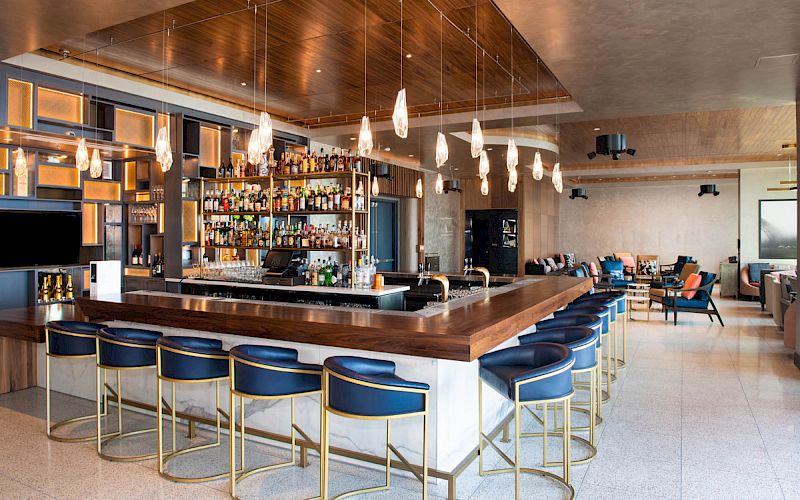 This image shows a modern bar interior with a long counter, blue barstools, hanging lights, fully stocked shelves, and seating areas in the background.