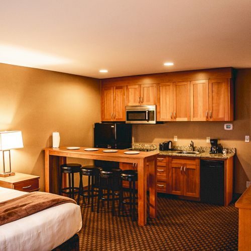 The image shows a hotel room with a bed, bedside table, lamp, kitchenette with a microwave, bar stools, TV, and a well-lit interior.