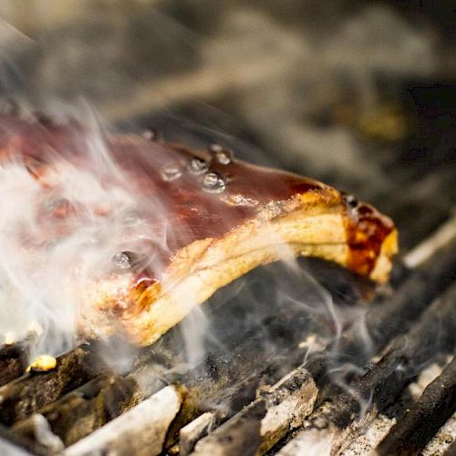 The image shows a piece of meat, possibly ribs, being grilled with smoky surroundings, and a layer of barbecue sauce and visible grill marks.