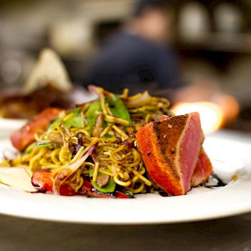 A plate of seared tuna, served atop a bed of mixed noodles and vegetables, garnished with sprouts and sauce.