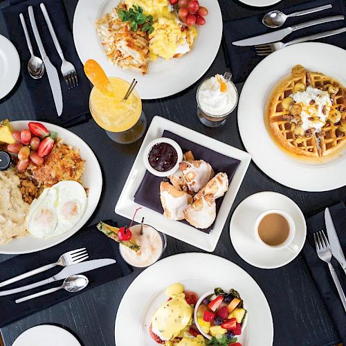 An overhead view of a breakfast spread, including eggs, waffles, pastries, fruit, pancakes, drinks, and coffee on a table with utensils and napkins.