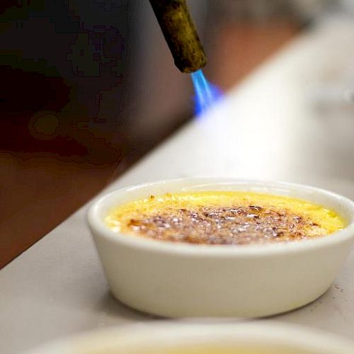 A culinary torch is caramelizing the sugar on top of a crème brûlée in a white ramekin.