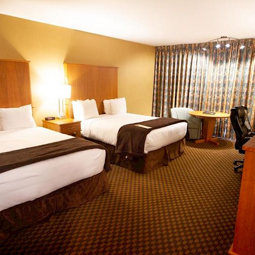 A hotel room with two double beds, a TV on a wooden dresser, a desk with a chair, and curtained windows. The beds have white linens and brown accents.