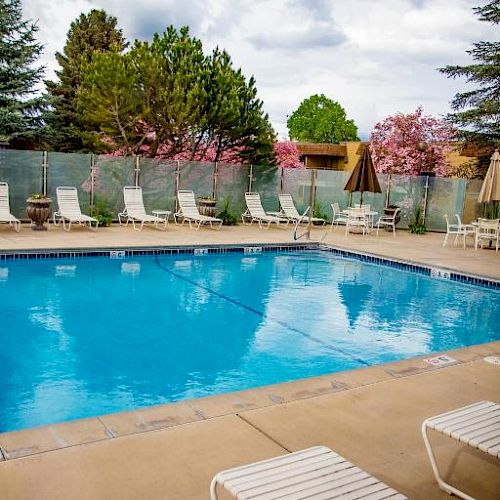 An outdoor swimming pool surrounded by lounge chairs, with trees and a fence in the background, under a partly cloudy sky.