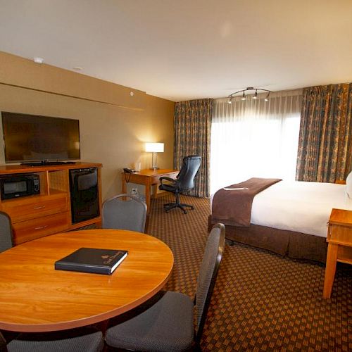 A well-lit hotel room with a single bed, table, chairs, TV, desk, and microwave, featuring curtains and carpeted floor, and a book on the table.