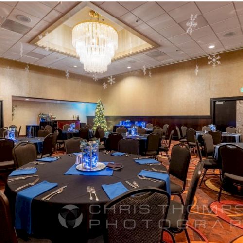 A decorated event hall with round tables, blue-themed centerpieces, and a chandelier, featuring a Christmas tree and snowflakes.