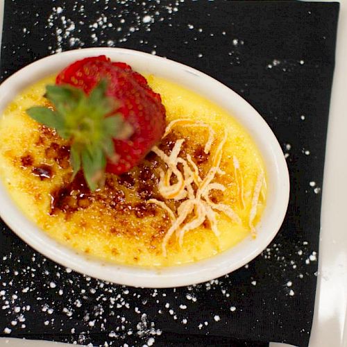 A dish of crème brûlée topped with a fresh strawberry and garnished with grated coconut, served on a black napkin with powdered sugar sprinkled around.