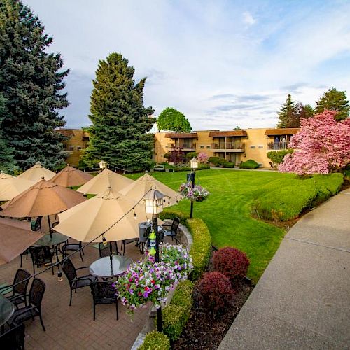 The image shows an outdoor seating area with tables and umbrellas, lush greenery, blooming trees, and a pathway leading to buildings in the background.