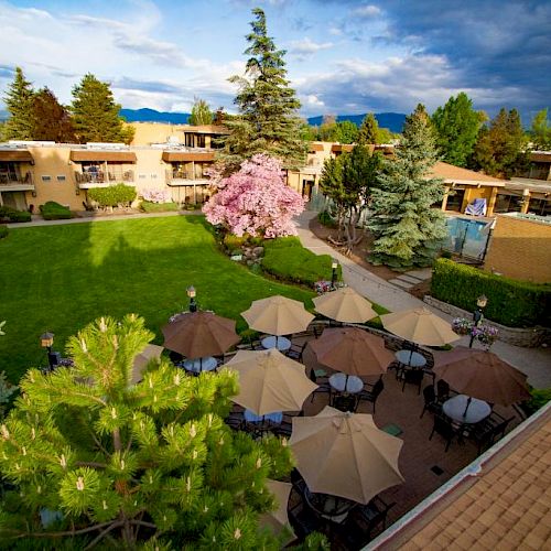 The image shows an outdoor courtyard with tables and umbrellas, surrounded by greenery and buildings under a partly cloudy sky.