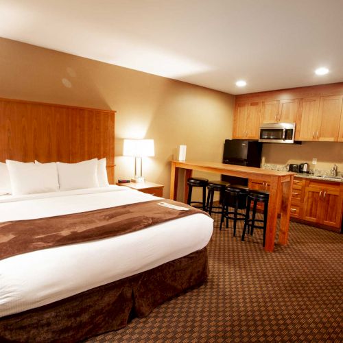 A cozy hotel room with a large bed, nightstands, lamps, and a kitchenette featuring a microwave, sink, and bar stools in a warmly lit environment.