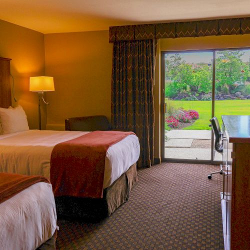 A hotel room with two beds, a TV on a dresser, a desk and chair, and a sliding glass door opening to a garden view outside.